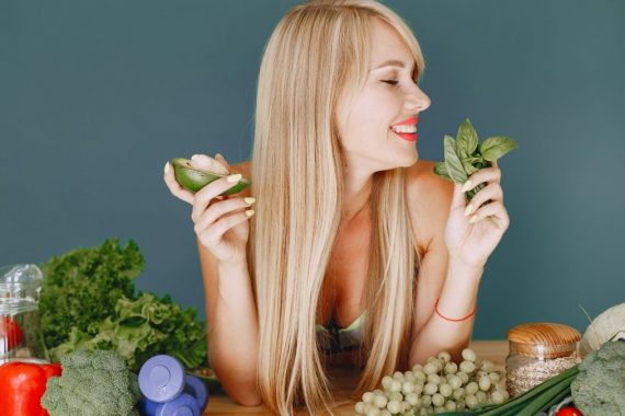 A smiling woman holding avocado and basil, surrounded by fresh vegetables and fruits for healthy eating.
