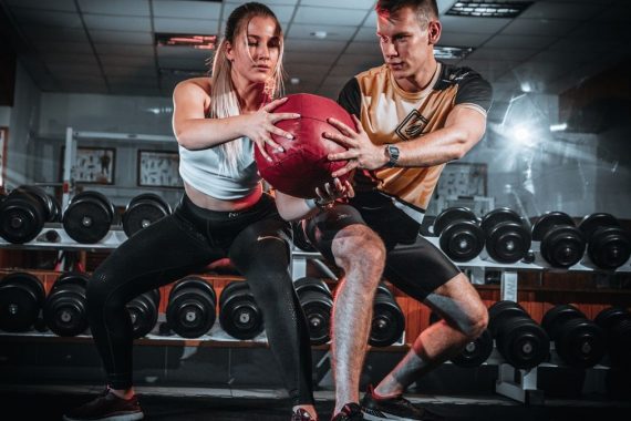 A man and woman perform a tandem workout with a medicine ball in a gym setting.
